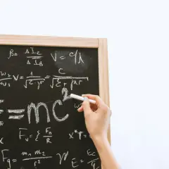 Tablica z równaniami fizycznymi, gdzie widać słynne E=mc². Ręka pisze kredą, a obok widnieje znak mniejsze lub równe.