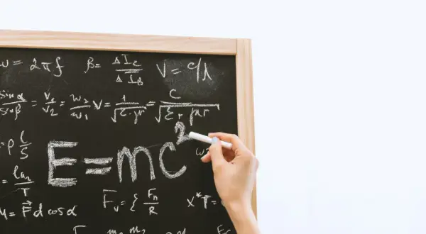 Tablica z równaniami fizycznymi, gdzie widać słynne E=mc². Ręka pisze kredą, a obok widnieje znak mniejsze lub równe.